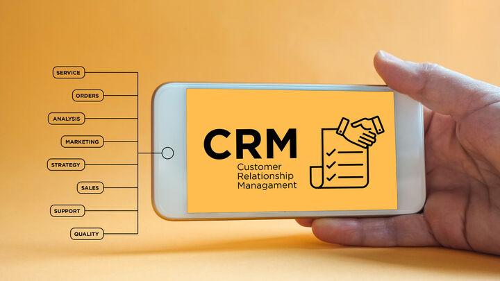 Hand holding smartphone displaying CRM (Customer Relationship Management) concept with business function icons including service, orders, analysis, marketing, strategy, sales, support, and quality