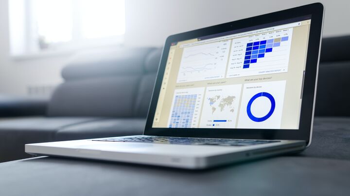 Laptop displaying analytics dashboard with blue data visualizations including charts, world map, and donut graph on a desk with blurred background.