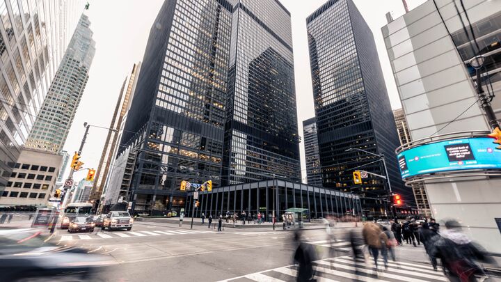 Wide-angle view of a downtown city intersection with tall black glass office towers housing financial institutions, pedestrians crossing in motion blur, cars waiting at traffic lights, and overhead streetcar wires on an overcast day.