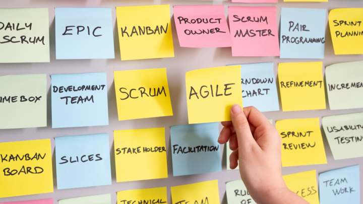 Hand placing sticky notes labeled with Agile methodology terms like 'AGILE', 'SCRUM', 'KANBAN', 'PRODUCT OWNER', and 'SCRUM MASTER' on a wall.