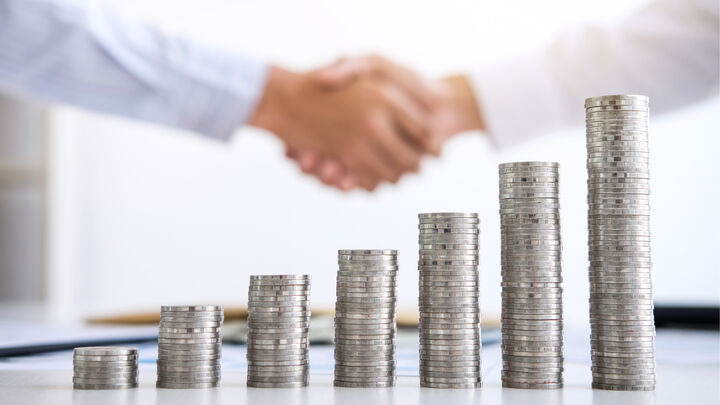 Business professionals shaking hands in background with ascending stacks of coins in foreground representing financial growth and partnership success.