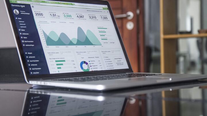 Laptop displaying analytics dashboard with graphs, charts, and data visualizations on glass desk in modern office setting