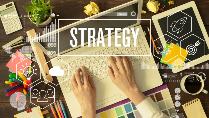 Overhead view of hands typing on a laptop with "STRATEGY" text overlay, surrounded by business planning items including a tablet, sticky notes, markers, coffee, and digital icons for rockets, gears, and targets.