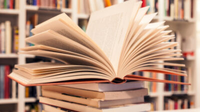 Open book with fanned pages resting on stack of closed books, with blurred bookshelf filled with colorful books in background