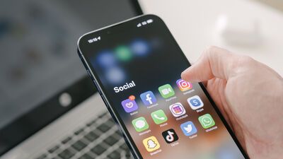 A man's hand holds a Iphone 13 Pro max with social media apps, against the background of a laptop.